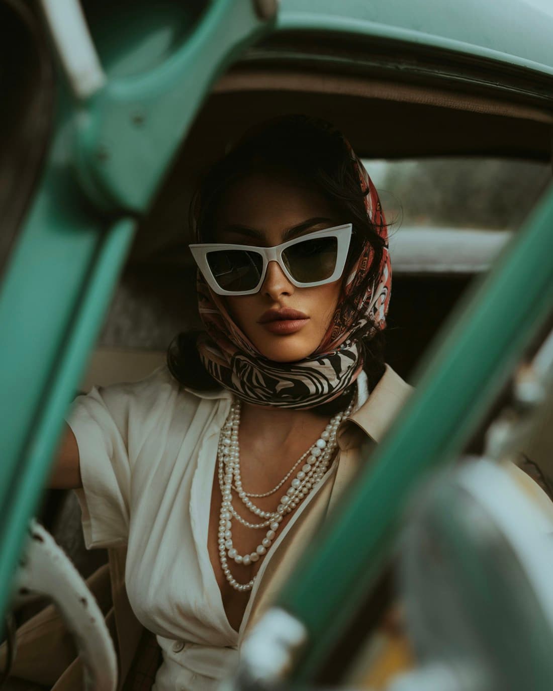 A young woman in white cat-eye See La Vie sunglasses, headscarf and pearls, seated in a vintage car